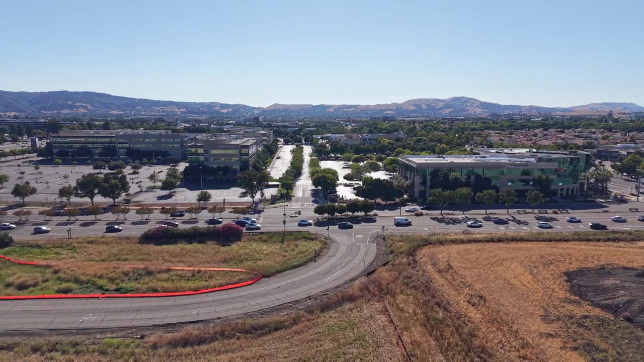 Dronevideo svæver over Dublin, Californien, afslører moderne kontorbygninger, brede gader, biler og gyldne marker under klar blå himmel. Klart dagslys fremhæver bydesign