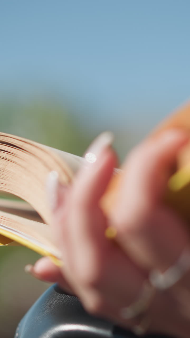 primer plano de la mano con uñas bien pulidas adornadas con anillos volviendo a través de las páginas del libro de portada amarilla contra un suave fondo borroso, mostrando un momento de lectura suave y reflexivo