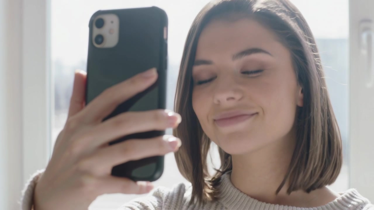 Una donna con un maglione di lana alza lo smartphone, si scatta un selfie vicino alla finestra, toccando il telefono e sorridendo.