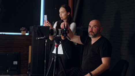 cameraman sitting behind the camera talks and gestures. caucasian girl standing at backdrop taking video on her phone.
