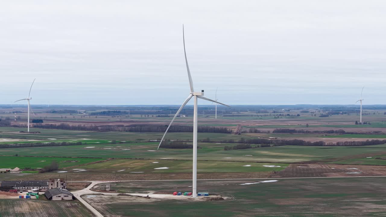 Vídeo com drone mostrando turbinas eólicas em meio a vastas terras agrícolas, destacando a energia renovável. A vista grande angular captura a paisagem natural e serena sob céus nublados.
