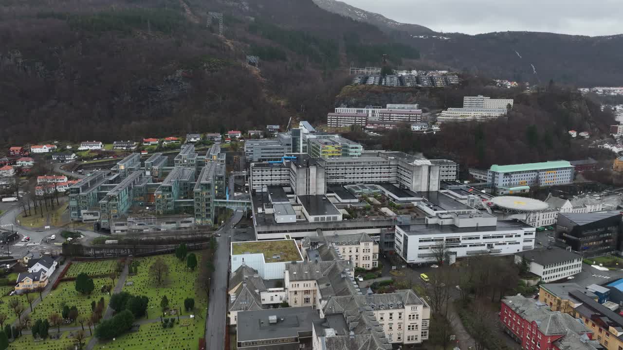 Haukeland Hospital in Bergen, Norway, with old and new buildings, graveyard, helicopter landing, and hospital exterior. Slow aerial ascent