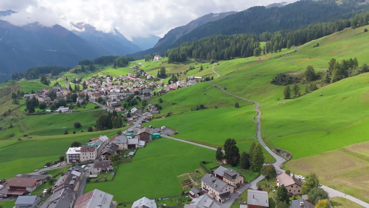 Aerial flyover Swiss town with houses and homes in idyllic alps landscape. Cloudy day in summer with meadows and snowy peaks of mountains. Tilt up wide shot