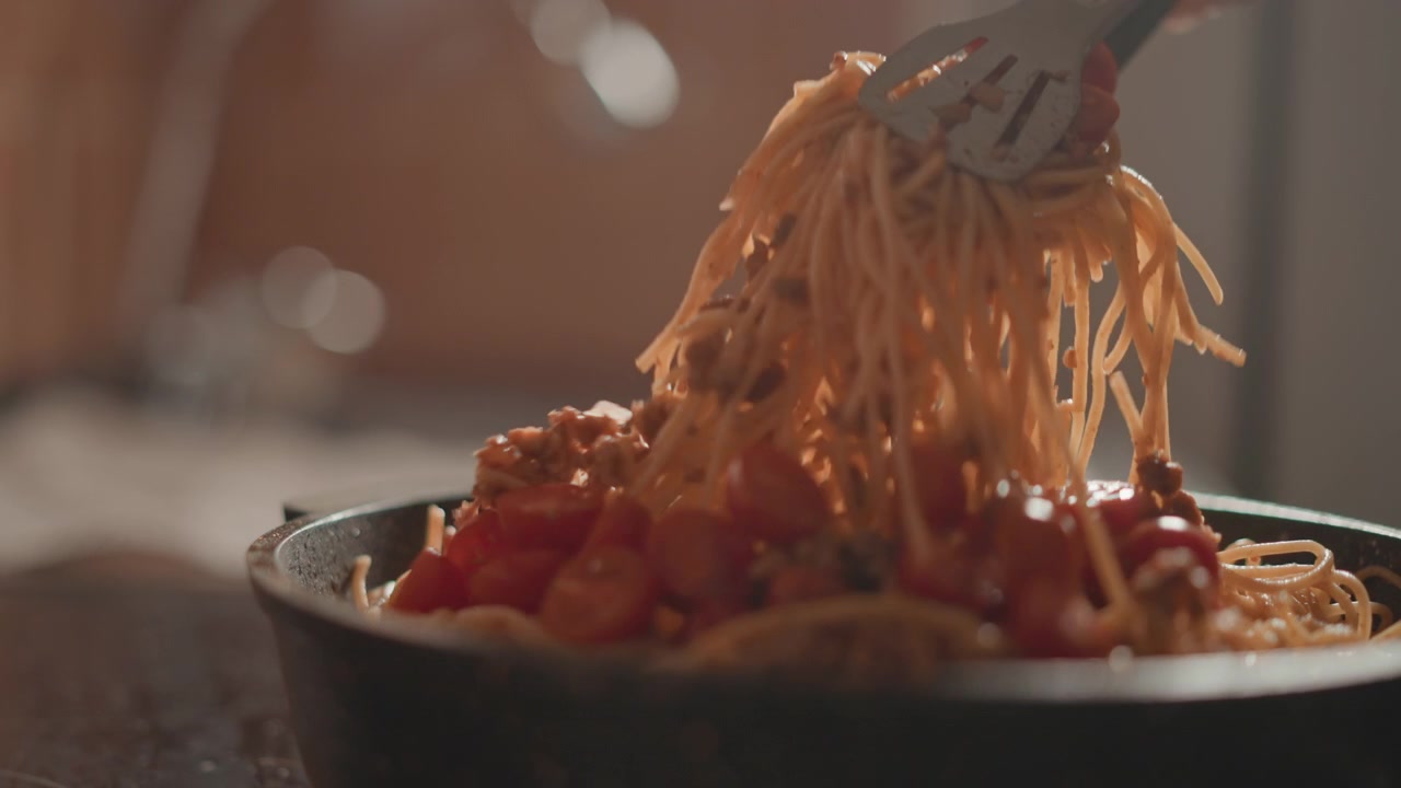 Scena di cucina dal basso con spaghetti sollevati e arrotolati da una padella bassa con pinze di metallo, salsa lucida che ricopre i noodles, pezzi di pomodoro sparsi, luce naturale calda e profondità di campo ridotta.