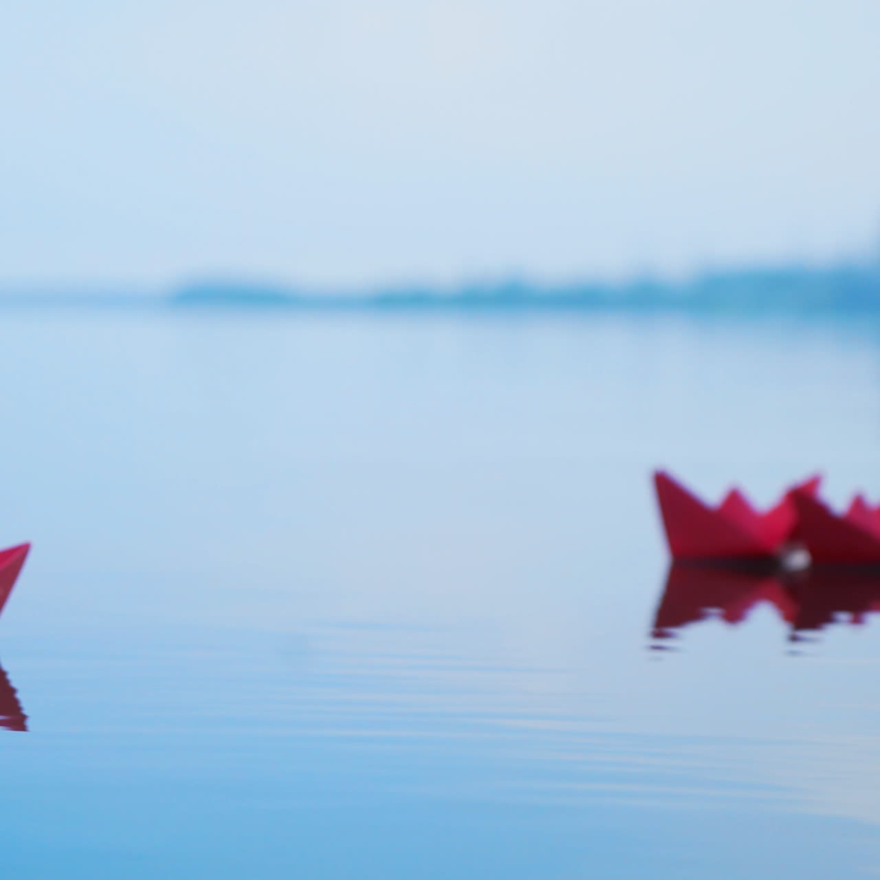 Red paper origami boats floating on the clear water with their reflection on blurred background. Three origami boats swimming on the blue lake. Close-up