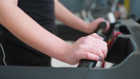 close up of white person wearing black outfit holding treadmill handle while exercising indoors with ash nail polish and blurred gym equipment in background during fitness workout session
