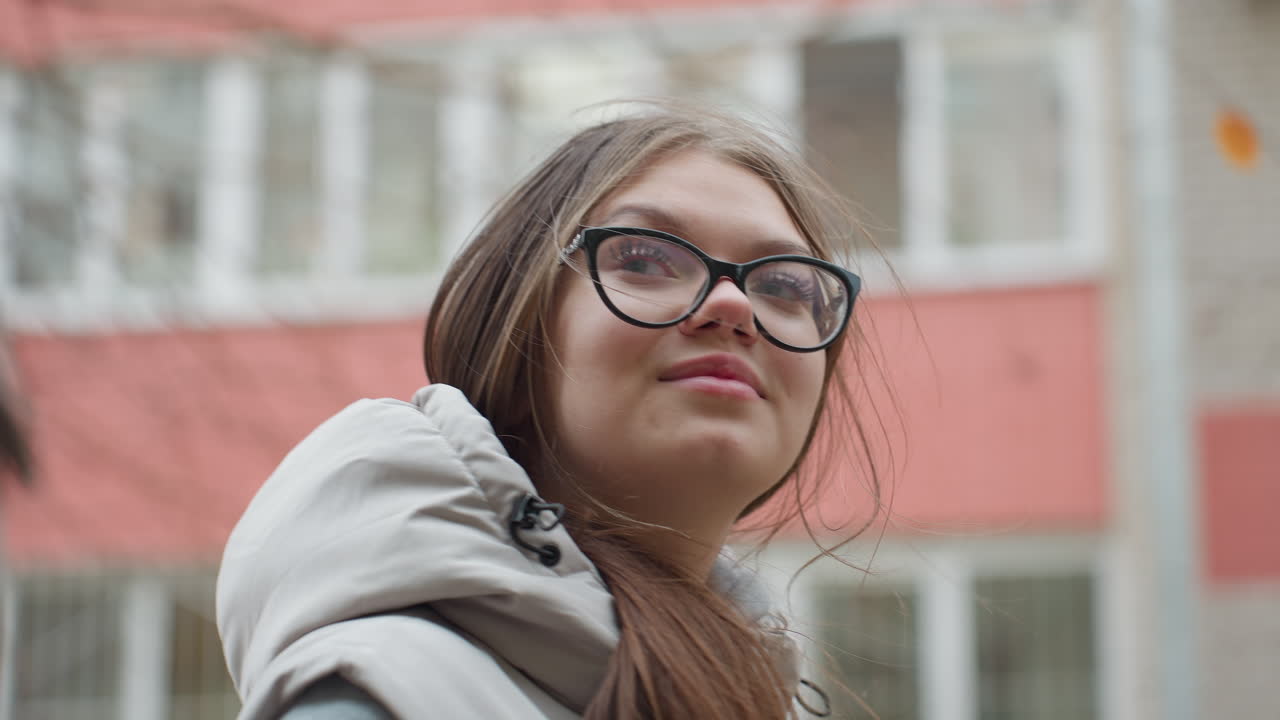 Close up of young lady in glasses looking at colleague with subtle smile as her hair sways with wind during outdoor stroll, dressed in warm jacket, background features blurred building