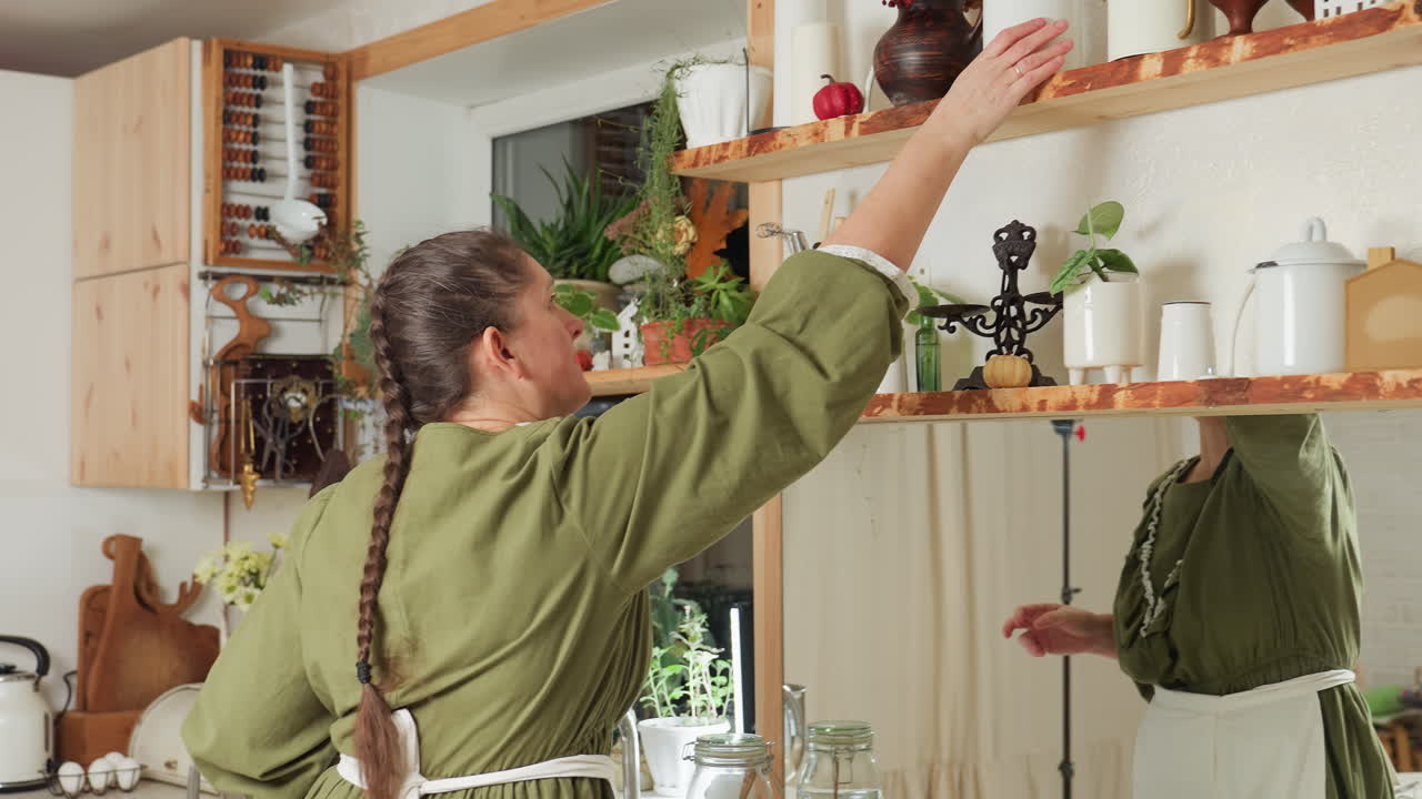 Chef arranges white glass jar back on wooden shelf in cozy kitchen with plants and utensils after using ingredients, while standing beside stainless steel bowl near sink, wearing green dress