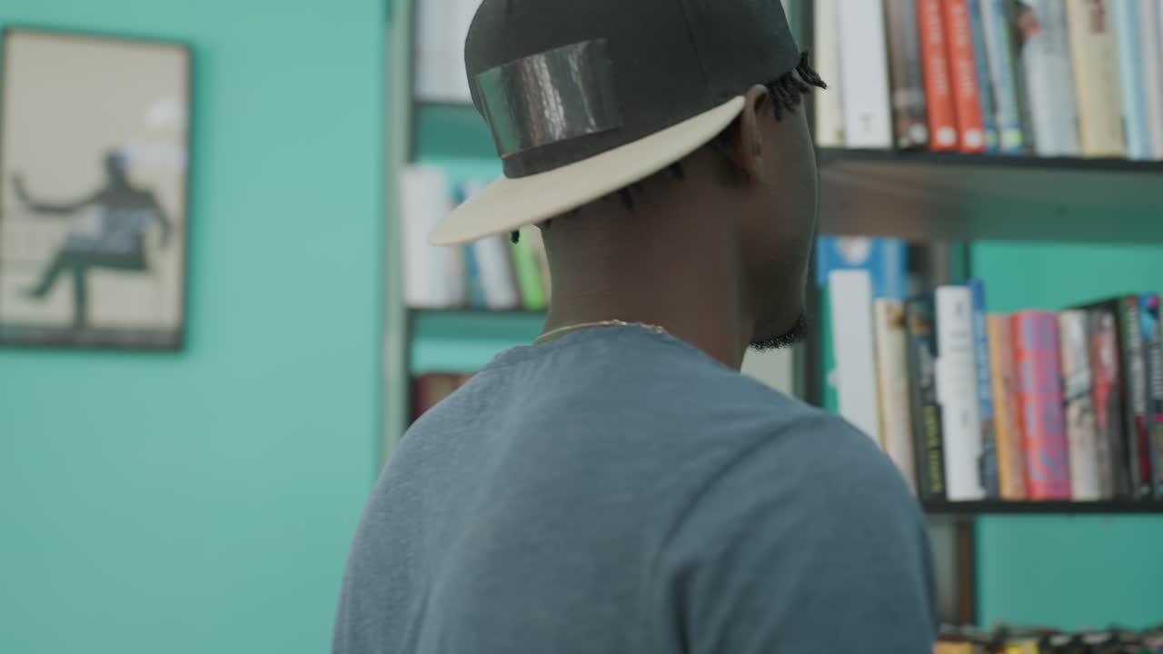 Student in casual cap studies shelves in library, side profile focused while scanning colorful spines near framed art on teal wall, sunlight from window softly fills quiet aisle