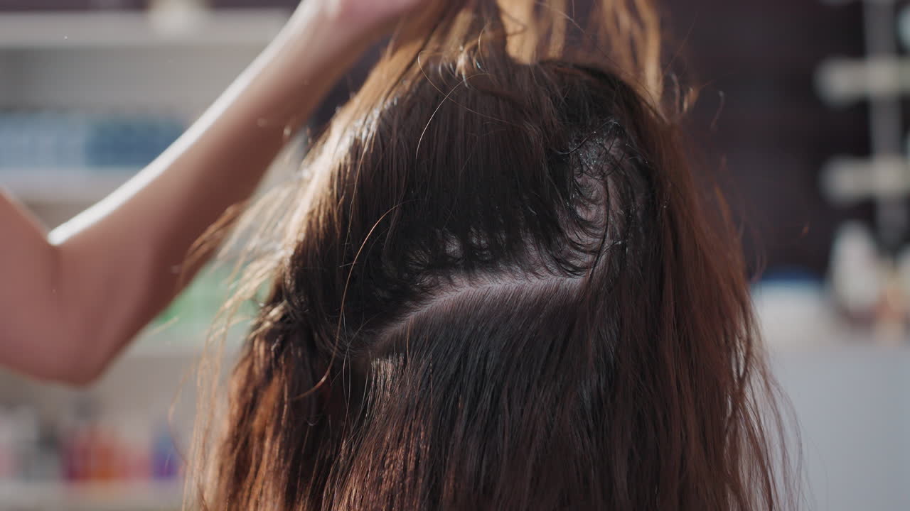 백인 컬러리스트가 윗부분에 뿌리 염색을 하고 있고, 안정적인 브러시 스트로크와 가르마 레이어, 고른 커버력과 깔끔한 가르마, 배경에는 컬러 제품 스튜디오 선반, 전문적인 기술