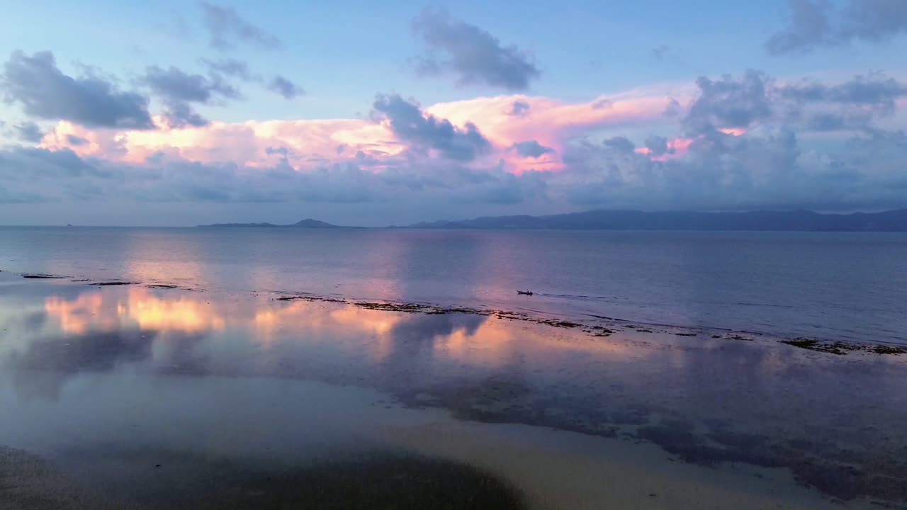 Drone footage of sunrise over Koh Phangan Thailand with a small boat moving across the calm tropical sea and the sunlight reflecting boat and clouds brightly on the water surface