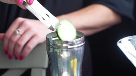 bartender drops and muddles mint leaves and cucumber slices in a silver cobbler shaker to make a cocktail using tongs and a muddler, closeup of woman’s hands with pink nail polish