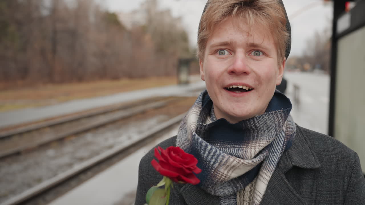Giovane uomo allegro che tiene una rosa sulla banchina del treno. Giovane uomo gioioso che sorride con un fiore in un paesaggio autunnale. Uomo sorridente e radioso presenta una rosa in una piacevole giornata autunnale tra pendolari indaffarati.