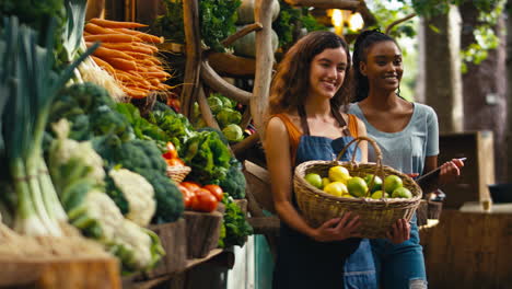 Retrato-De-Dos-Mujeres-Sonrientes-Con-Tableta-Digital-Trabajando-En-Un-Puesto-De-Frutas-Y-Verduras-Frescas-En-El-Mercado