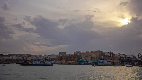 Timelapse-Frente-A-La-Costa-Del-Romántico-Pueblo-Pesquero-De-Marsaxlokk-En-La-Isla-De-Vacaciones-De-Malta-Con-Vistas-A-Los-Barcos-De-Carga-Flotantes-Y-Barcos-De-Pesca,-Así-Como-A-Los-Edificios-Históricos.