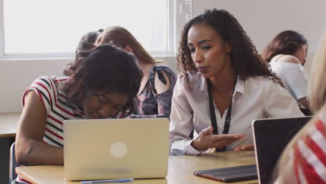 Woman-Teacher-Giving-One-To-One-Support-To-Female-Student-Working-At-Desk-On-Laptop