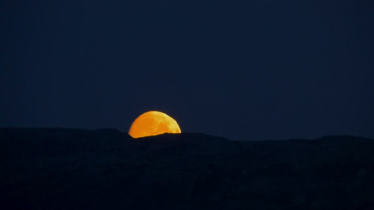 CLOSE UP view of the moon glowing reddish orange as it sets behind a distant mountain ridge