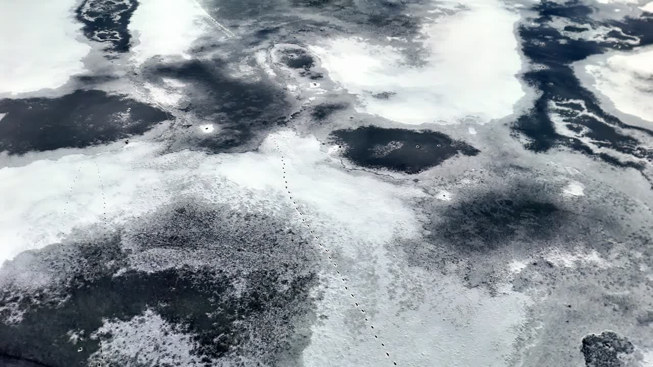 A frozen lake displays dark ice patches and a dotted trail crossing its textured snow-covered surface, captured from above in a cold and remote winter landscape.