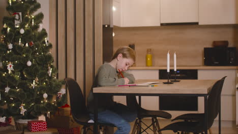 side view of a girl in green sweater writing a letter of wishes sitting at a table in a room decorated with a christmas tree