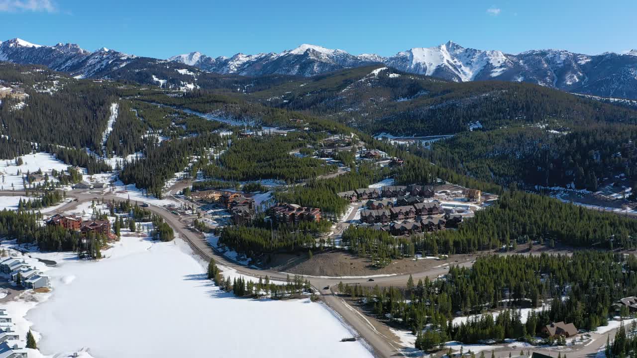 Aerial footage shows winter condos in Big Sky, Montana, dusted in snow and nestled among tall pines, with quiet streets and panoramic mountain backdrops in a calm, scenic town setting
