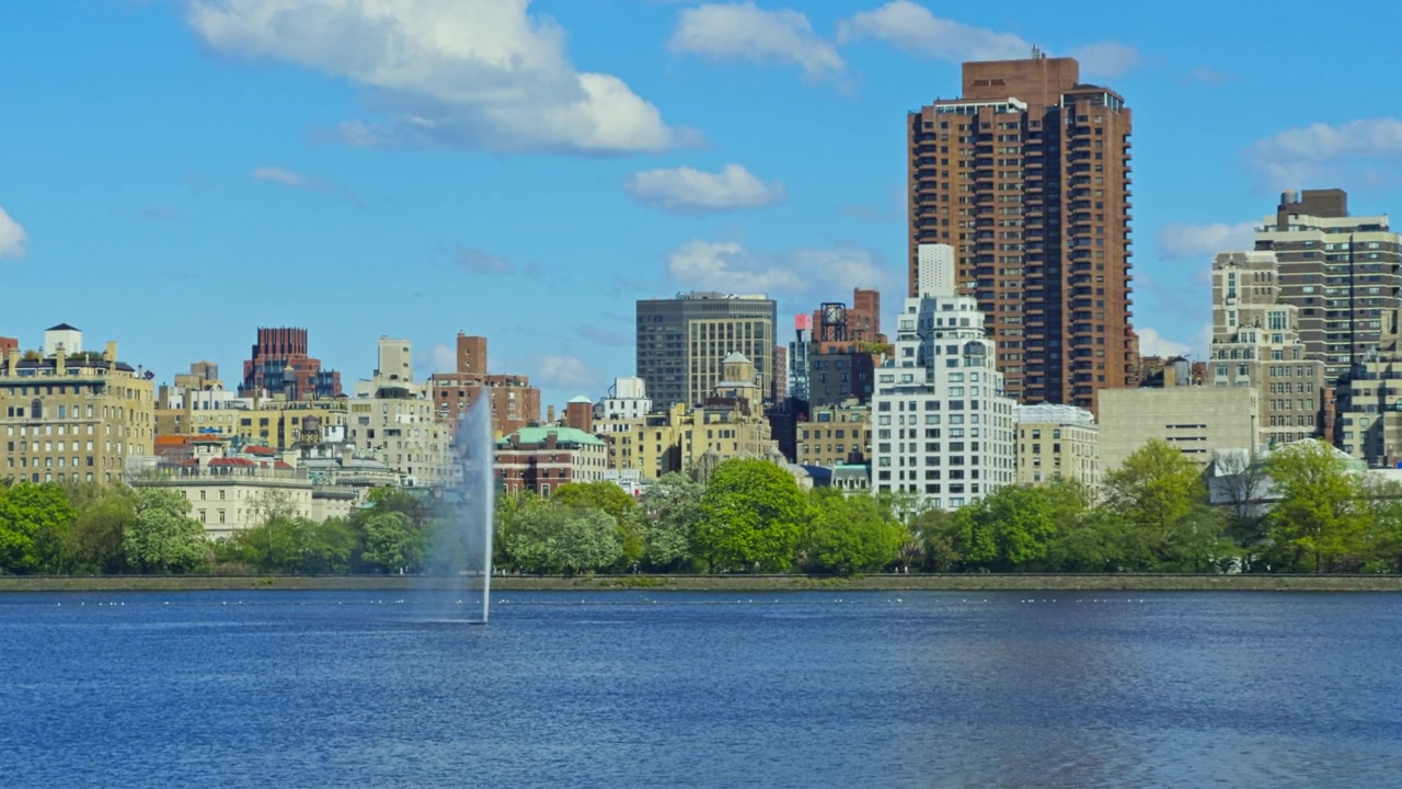 Edifícios de Manhattan elevando-se além da fonte do Reservatório Onassis no Central Park, à luz do dia.