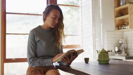 Glückliche-Kaukasische-Frau-Sitzt-In-Der-Cottage-Küche-Und-Liest-Ein-Buch,-Mit-Teekanne-Auf-Der-Theke