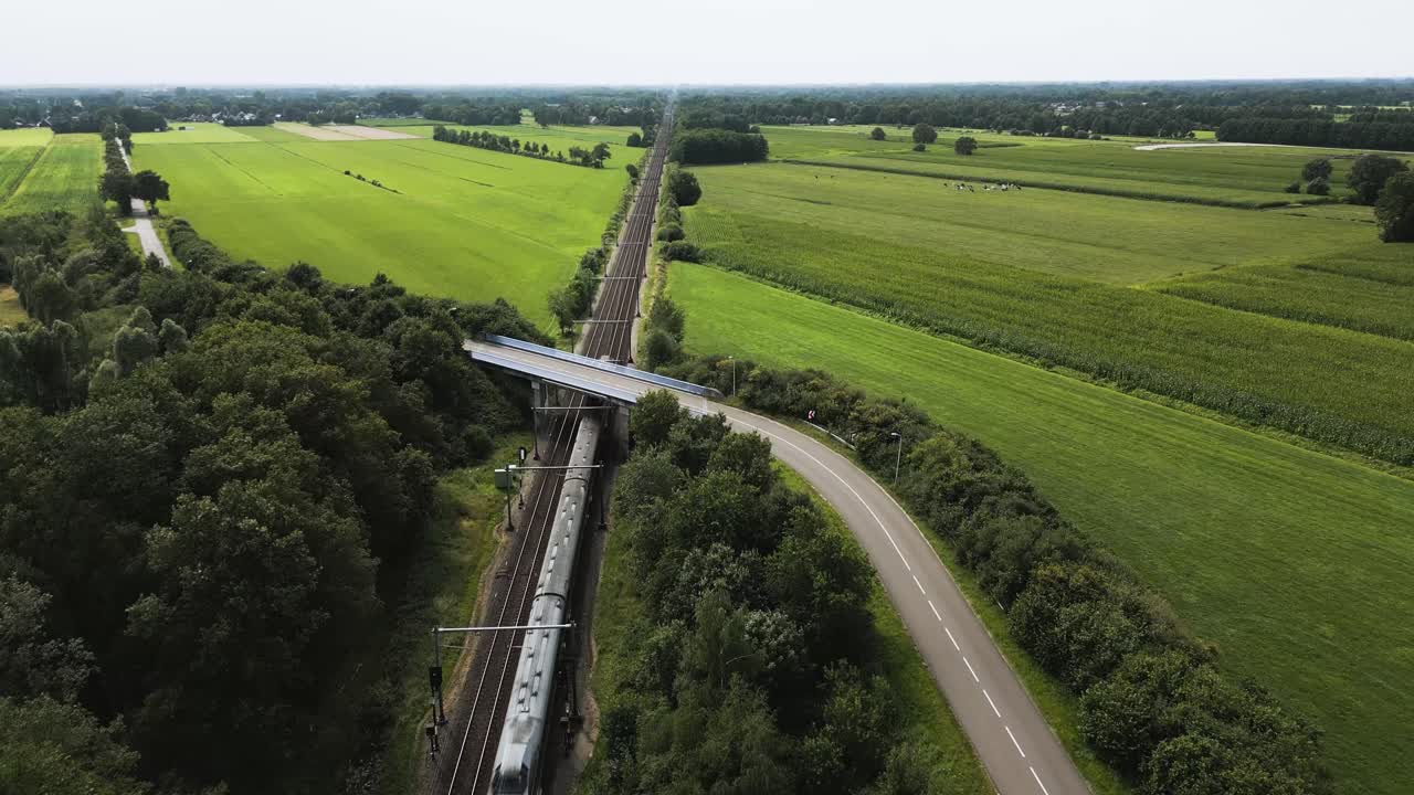 Aerial view of passenger train moving through lush green farmlands and countryside road in Twente region showing modern transport network blending with peaceful rural Dutch landscape