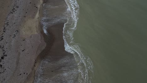 overcast day on a uk shingle beach, viewed from directly above. gentle waves wash the textured shoreline. a serene, top-down perspective of the british coast.