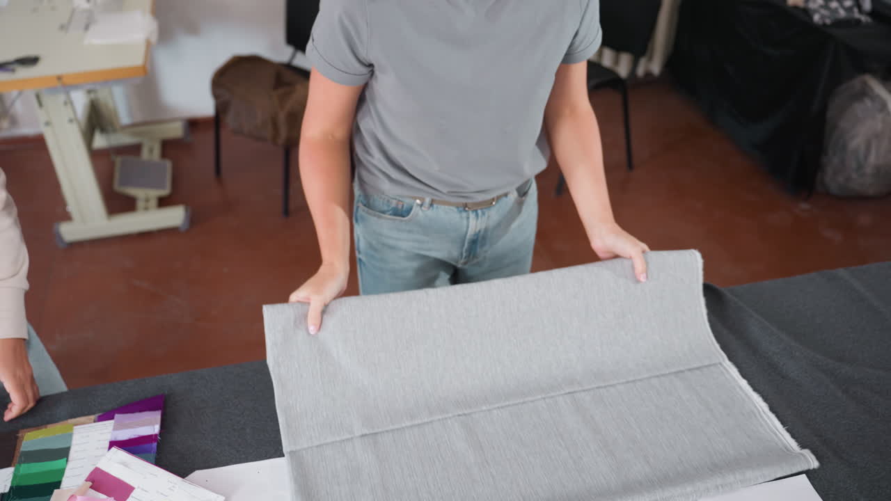 Aerial view of fashion design trainer guiding lady on fabric folding technique in bright workspace, hands carefully adjusting soft gray textile surrounded by colorful swatches, and creative materials