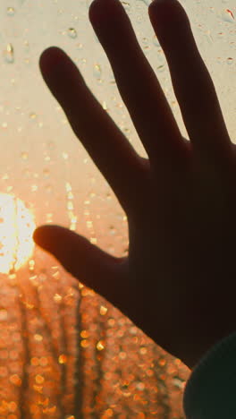 silhouette of hand reaching to red sun through wet window closeup. hand touches glass with clear raindrops at dusk on rainy day. emotional state