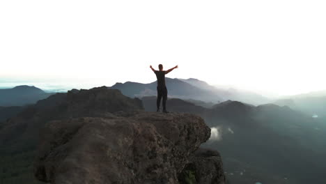 hombre encima del roque nublo en gran canaria estirando los brazos y disfrutando de la vista mientras el dron hace un vuelo orbital al atardecer mientras la niebla está sobre las rocas entre los bosques