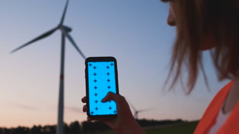 woman engineer holding a smartphone with chroma key and making scroll down gesture on the background of wind turbine at sunset.