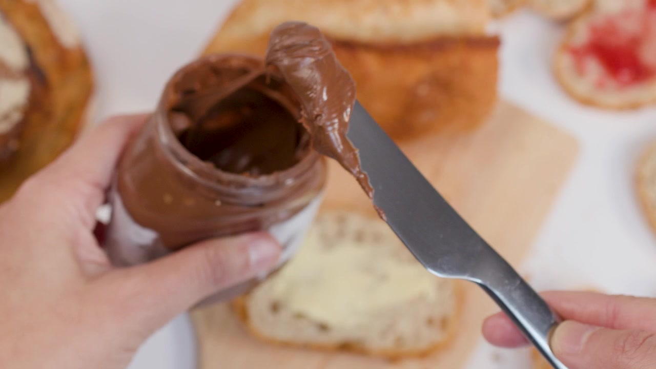 Primo piano di mani che usano un coltello per spalmare crema al cioccolato da un barattolo sul pane
