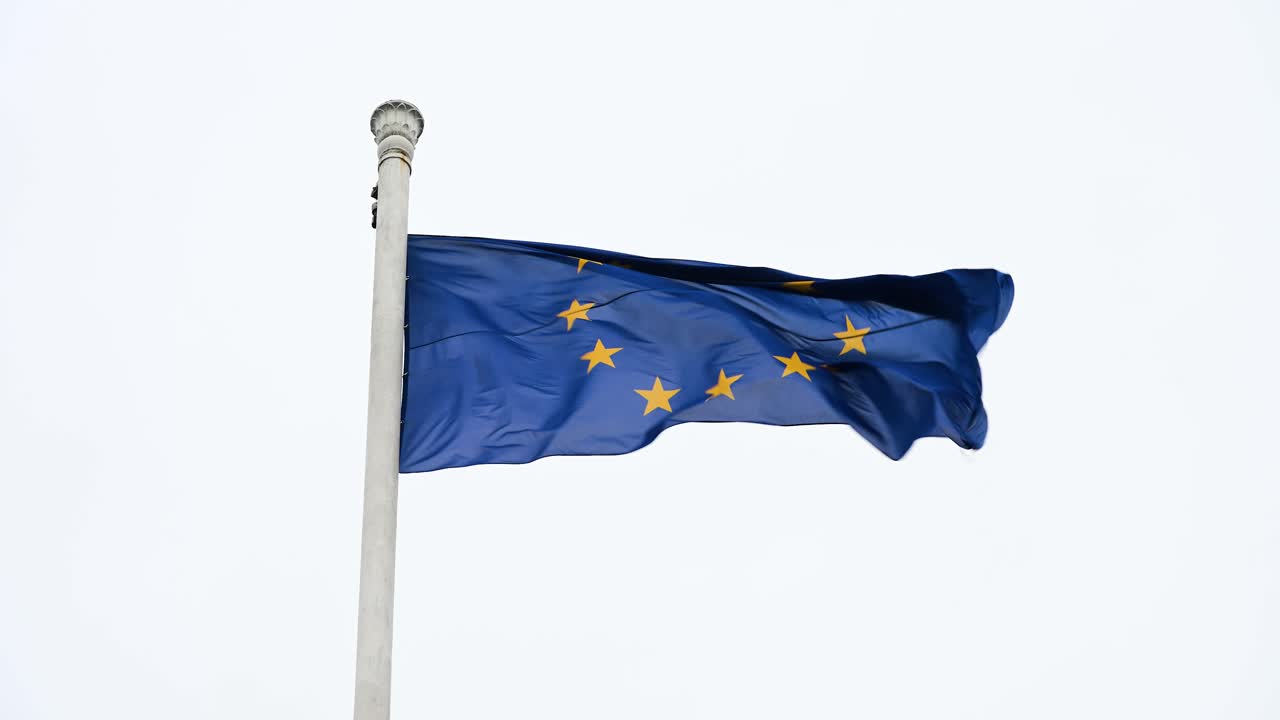 Ripresa al rallentatore della bandiera dell'Unione Europea (UE) che sventola su un'asta alta, spazzata dal vento in una giornata nuvolosa, enfatizzando il movimento fluido della bandiera mentre ondeggia con la brezza.