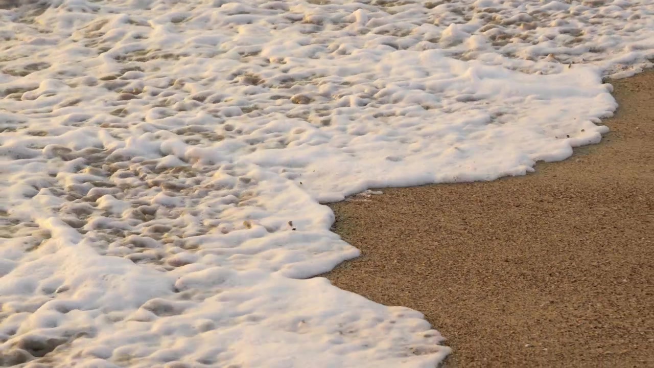 Slowmotion silkebløde havbølger og hvidt havskum, der skyller ind over en sandstrand på en tropisk kystlinje under en smuk gylden solnedgang