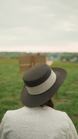 a scene capturing the back view of a woman wearing a stylish hat and a white dress, seated on a chair in a lush grass field beside a tranquil lake. a large drawing board is visible in the background