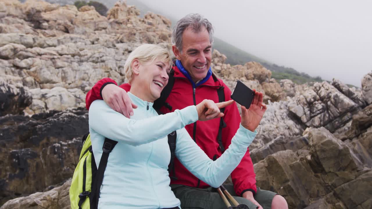 pareja de excursionistas mayores con mochilas sentados en las rocas y sonriendo mientras tienen una videollamada