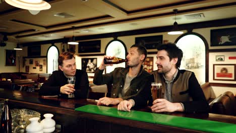 three young men in casual clothes are drinking beer in fancy sports bar, clanging glasses and bottles and talking while sitting at counter. guys having night out concept.