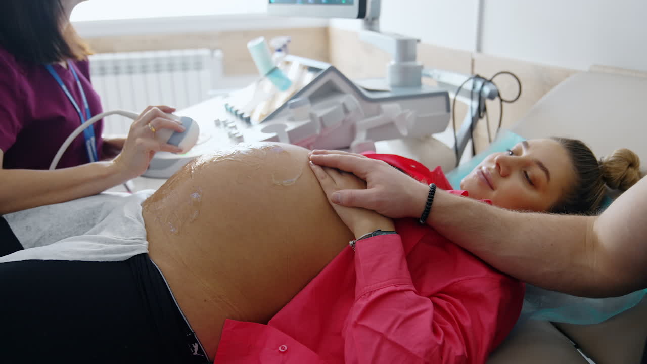 Donna caucasica incinta si sottopone a un'ecografia. Una mano maschile le tiene la mano per darle supporto.