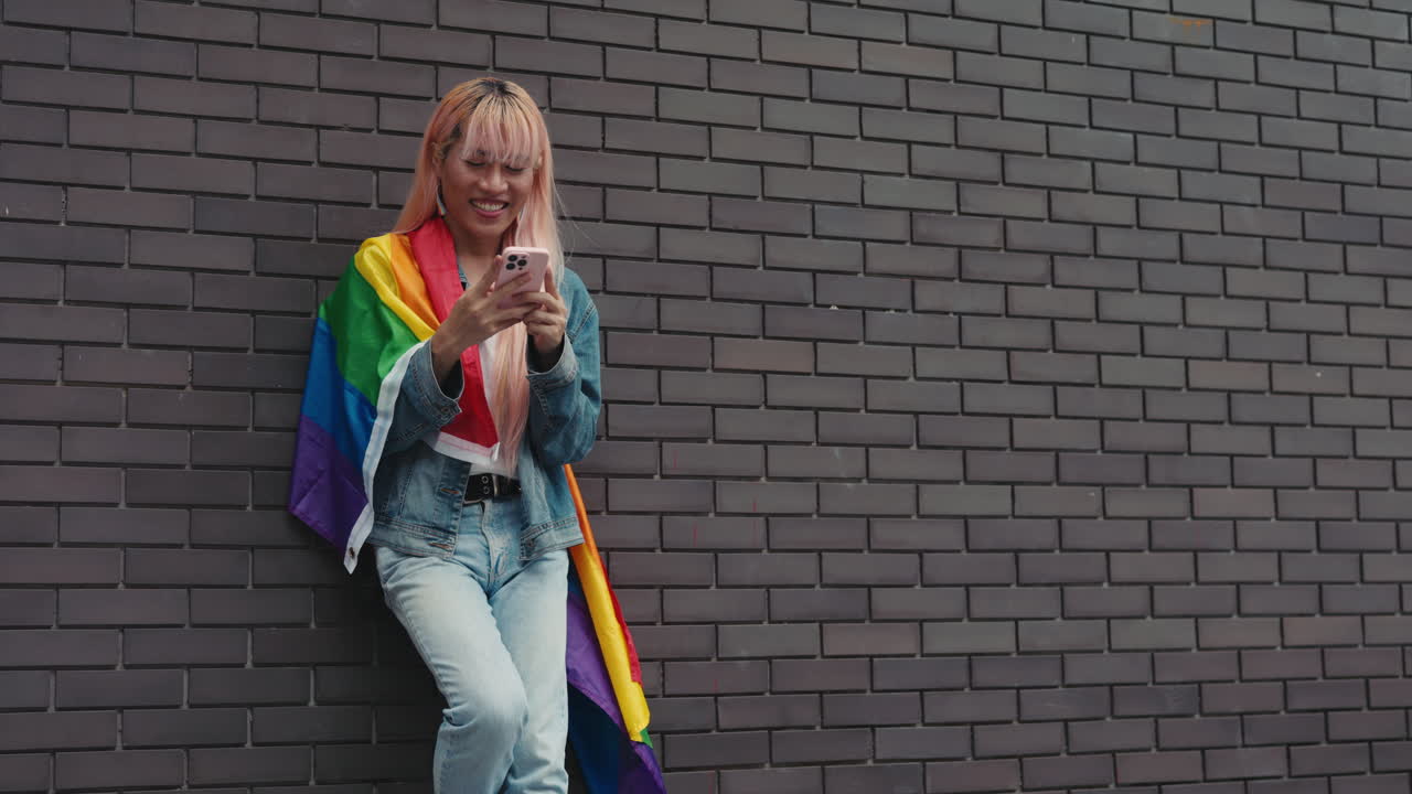 Woman using phone while holding rainbow flag leaning against a brick wall