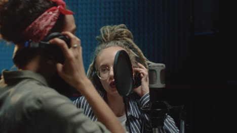 close up of the two happy young attractive female singers recording a duet song while singing in the mic in the dark sound studio