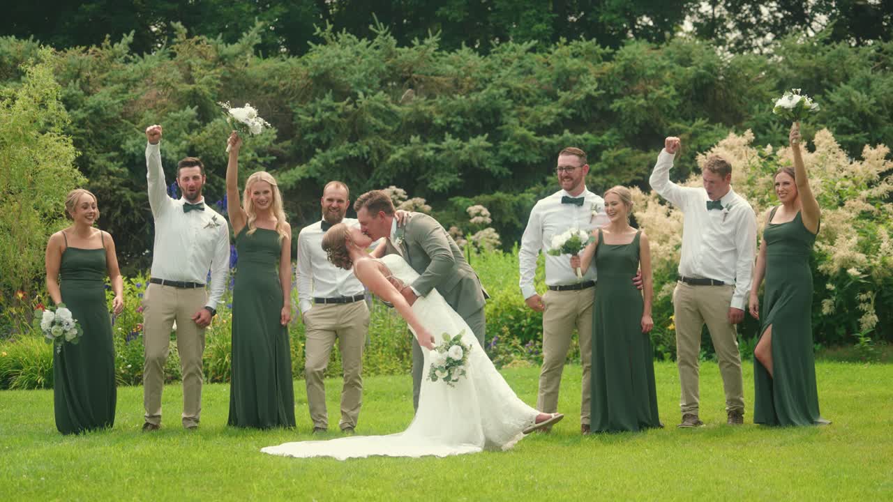 Groom dips and kisses bride in a garden while their bridal party cheers in celebration