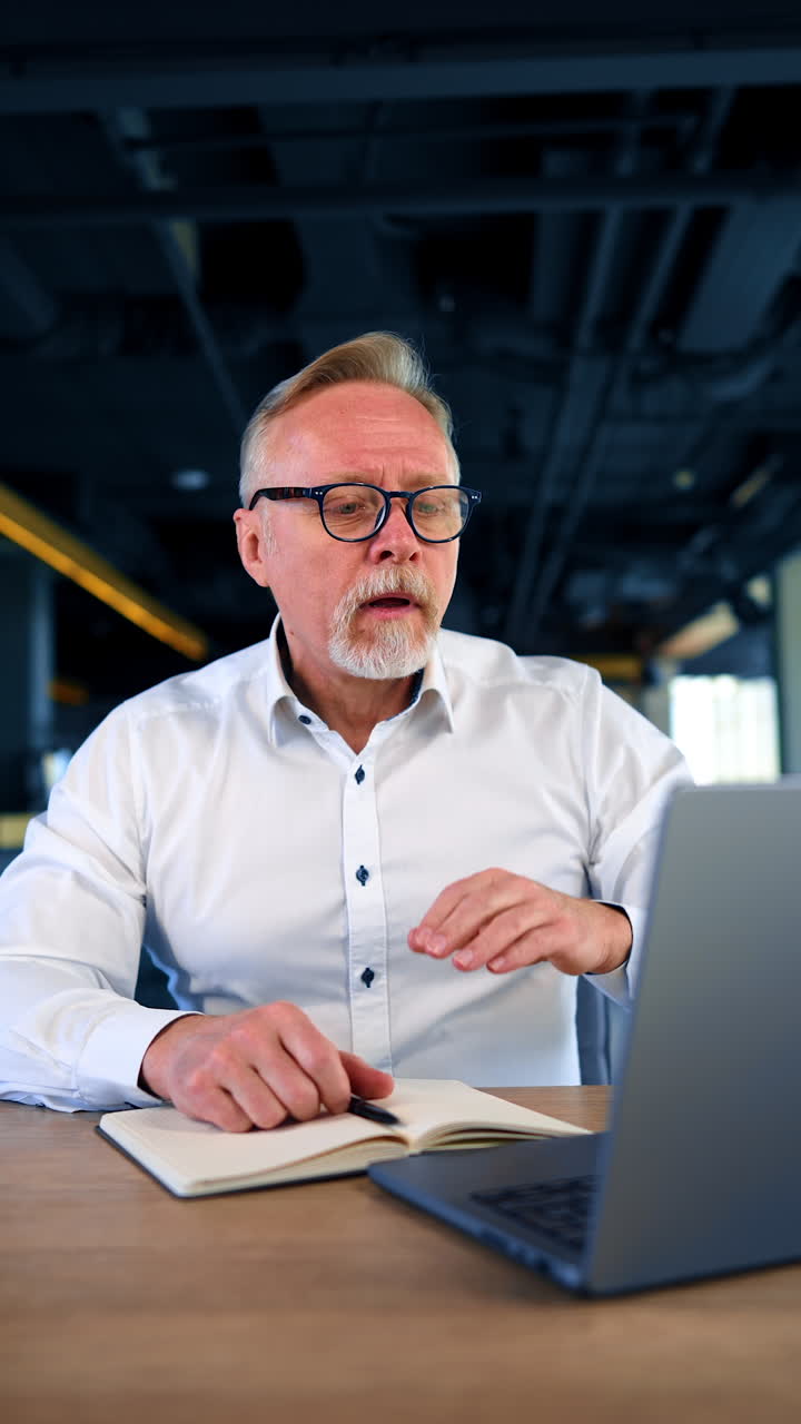 Puzzled adult man sitting at desk looks closely at his laptop. Man gestures rudely to the computer screen. Vertical video.