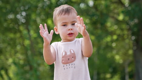 beautiful toddler says something gesturing with hands. kid waves his hands and looks at camera. blurred greenery of nature at backdrop.