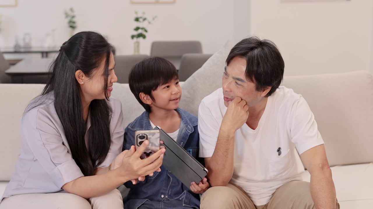 Happy Asian family bonding on sofa, using smartphone and tablet in bright, cozy living room