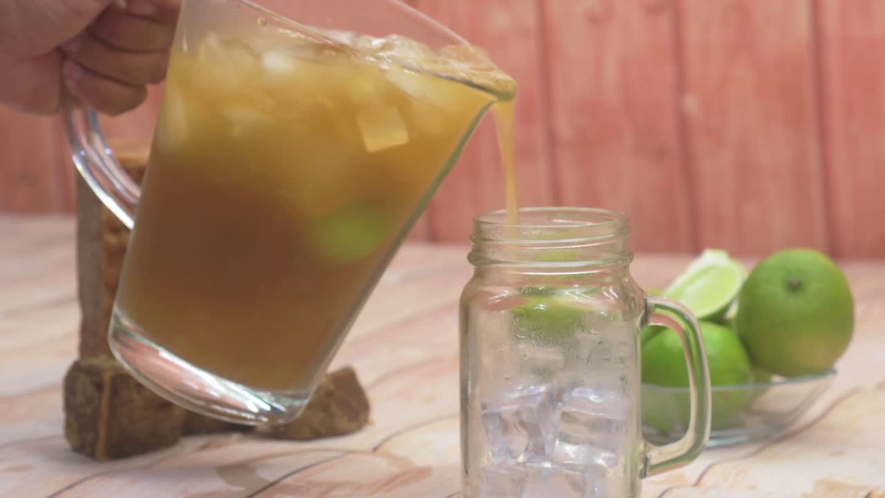 Refreshing close-up hand preparing to pour homemade Papelón con Limón into a glass mason jar filled with ice, amidst fresh limes and raw sugar chunks