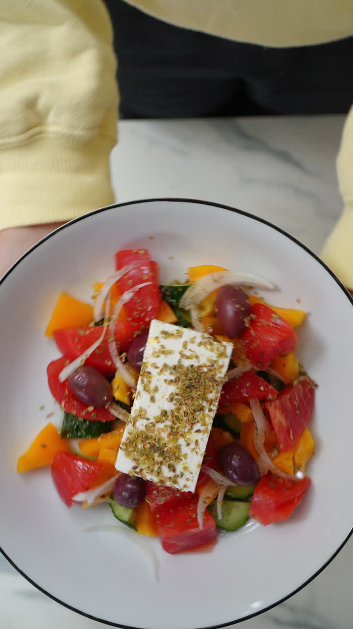 Vertical footage Hands of a chef presenting a plate of fresh greek salad with feta cheese, tomatoes, cucumber, olives and onions, seasoned with oregano