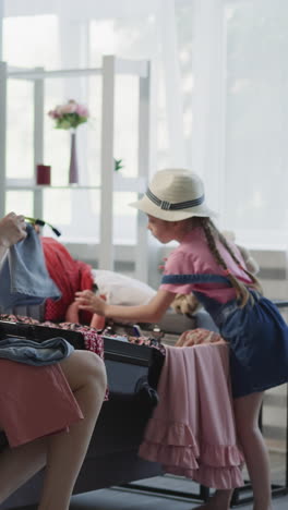 mother gathers apparel while preschooler daughter puts doll and teddy bear into suitcase in living room. woman and girl prepare items for travel