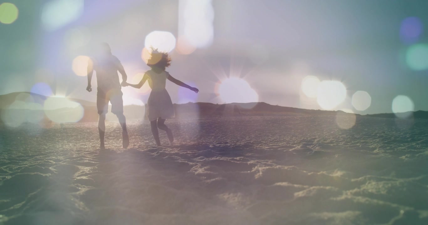 Couple se tenant la main marchant sur le sable vers l'horizon, le soleil créant un effet de flou artistique (bokeh) en superposition pour le marketing