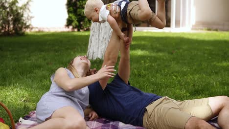Young-couple-with-a-baby-resting-in-the-park-on-the-grass.-Happy-family-of-three,-with-a-baby,-blonde-haired-boy.-Beautiful-parents-mom-and-dad-lying-on-a-plaid.-Dad-is-holding-child-with-his-hands